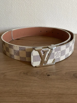 Rutig -bälte i beige - Snyggt bälte med klassiskt rutigt mönster i beige och vitt. Bältet har en stor  metal silver som spänne och insidan är ljusbrun. Perfekt accessoar för att lyfta din outfit.