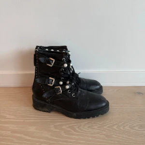 Svarta boots med pärldetaljer Zara - Tuffa svarta kängor med snörning, tre spännen och dekorativa pärlor. Grov sula och dragkedja baktill för enkel på- och avtagning. Perfekta för dig som vill ha en edgy stil med coola detaljer.