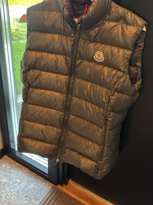 Mörkbrun dunväst från Moncler - Snygg mörkbrun dunväst från Moncler med hög krage och dragkedja framtill. Västen har ett quiltat mönster och Moncler-logga på bröstet. Perfekt för lager-på-lager och håller dig varm under kyliga dagar.