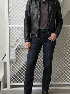 Svart klassisk skinnjacka - Snygg svart skinnjacka med raka linjer och ståkrage. Jackan har dragkedja framtill, två bröstfickor med dragkedja och knäppning vid midjan. Perfekt för en cool och tidlös look. En trasig zipper på bilden 