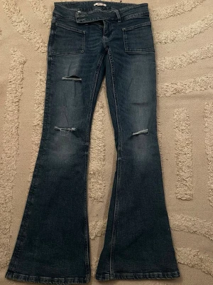 Blå utsvängda jeans med slitningar - Säljer ett par blå jeans med utsvängda ben och slitna detaljer på framsidan. Jeansen har låg midja, stora framfickor och klassiska bakfickor. Materialet är denim och färgen är tvättad blå. Perfekta för dig som gillar retrostil och trendiga detaljer.