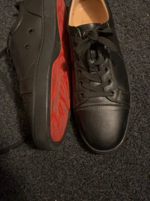 Svarta sneakers med röd sula - Snygga svarta sneakers i läder med klassisk låg profil och svarta skosnören. Skorna har en unik röd sula med grafiskt mönster undertill som sticker ut. Perfekta för dig som vill ha en stilren men ändå iögonfallande look.