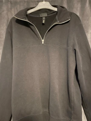 Svart hoodie med halv dragkedja - En svart hoodie med halv dragkedja framtill och hög krage. Tillverkad i mjukt sweatshirtmaterial och har långa ärmar. Enkel och stilren design som passar till det mesta.
