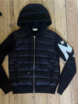 Svart pufferjacka från Moncler - Snygg svart pufferjacka från Moncler med huva och dragkedja framtill. Jackan har stickade ärmar och detaljer i vitt på ena ärmen samt Moncler-logga. Perfekt för dig som vill ha en sportig och trendig look. Materialmix med dun och bomull.