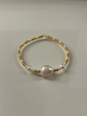 Pärlarmband med gulddetaljer - Snyggt armband med små vita och guldiga pärlor samt en större vit pärla i mitten. Armbandet har en elastisk tråd som gör det enkelt att ta på och av. Perfekt accessoar för att lyfta din stil.