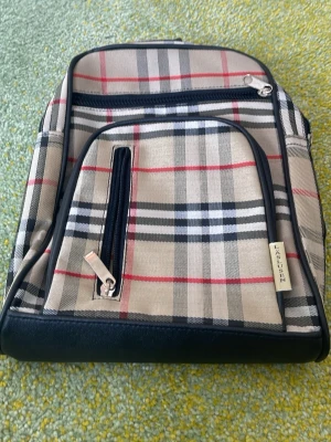 Rutigt ryggsäck med svarta detaljer - Snygg ryggsäck med klassiskt beige, svart och rött rutmönster. Väskan har svarta detaljer i konstläder, flera dragkedjefack och justerbara axelband. Perfekt för dig som vill ha en stilren och trendig accessoar till skolan eller stan.