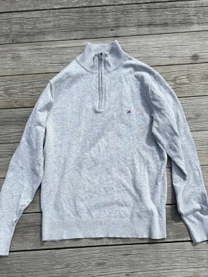 Ljusgrå halvzip från Tommy Hilfiger - Ljusgrå stickad tröja från Tommy Hilfiger med kvartszip och liten broderad logga på bröstet. Tillverkad i mjuk bomull, perfekt för lager-på-lager. Klassisk och stilren design med ribbade muddar och krage.