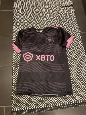 Svart Inter Miami Messi matchtröja - Säljer en svart Inter Miami matchtröja med rosa detaljer och tryck. Framsidan har klubbmärke och sponsorlogga, baksidan har Messi och nummer 10 i rosa. Tillverkad i lätt och ventilerande material, perfekt för fotboll eller träning.