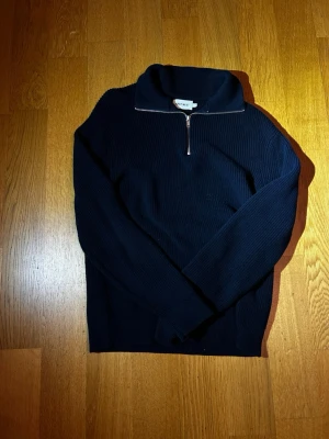 Mörkblå stickad halvzip tröja - Snygg mörkblå stickad tröja från East West med halvzip och hög krage. Tröjan har ribbad struktur och långa ärmar, perfekt för lager på lager. Passar dig som gillar en enkel men stilren look.                          Axelbredden=50cm. Längden=70cm