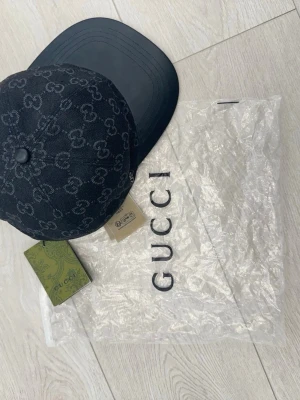 Svart keps från Gucci med monogram - Snygg svart keps från Gucci med klassiskt GG-monogram över hela kepsen. Skärmen är i svart och materialet ser ut att vara en mix av textil och läder. Perfekt accessoar för en trendig streetstyle-look.