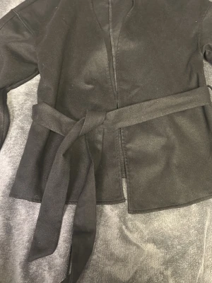 Svart kappa med knytskärp - Stilren svart kappa med knytskärp i midjan och rak passform. Kappan har långa ärmar och är tillverkad i ett mjukt tyg som ger en clean look. Perfekt för dig som gillar minimalistisk stil och vill ha en enkel men snygg jacka till hösten eller våren.☺️