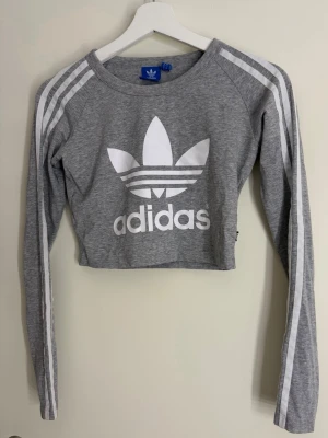 Adidas tröjor set - Säljer en grå croppad långärmad topp från Adidas med klassiska vita ränder längs ärmarna och stort vitt Adidas-tryck framtill. Tillverkad i mjuk bomullsmix, perfekt för en sportig och trendig look. Det går att köpa separata också :)