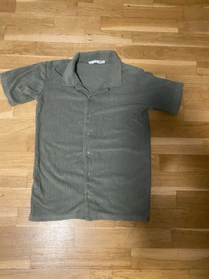 Grön ribbad kortärmad skjorta - Kortärmad skjorta i en dämpad grön färg med ribbad struktur och knappar framtill. Skjortan har klassisk krage och är tillverkad i ett mjukt material som påminner om manchester. Perfekt för en avslappnad stil.