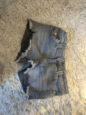 Grå jeansshorts med fransar - Säljer ett par grå jeansshorts med fransiga kanter och slitna detaljer. Shortsen har fickor både fram och bak, bälteshällor och knappstängning. Perfekta för varma dagar och en avslappnad stil.