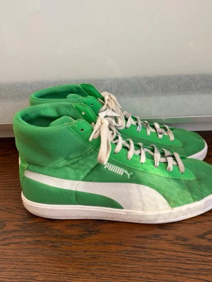 Gröna höga sneakers från Puma - Snygga gröna höga sneakers från Puma med vit sula och vita detaljer på sidan. Skorna har vita skosnören och är tillverkade i canvasmaterial. Klassisk Puma-logga på sidan och plös. Perfekta för en sportig och avslappnad stil. 42/42,5