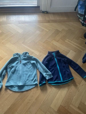 Två fleecejackor med dragkedja - Säljer två fleecejackor i olika färger, en ljusblå och en mörkblå med turkosa detaljer. Båda har hel dragkedja framtill, hög krage och långa ärmar. Perfekta för lager-på-lager och sköna att ha på sig under kyliga dagar.