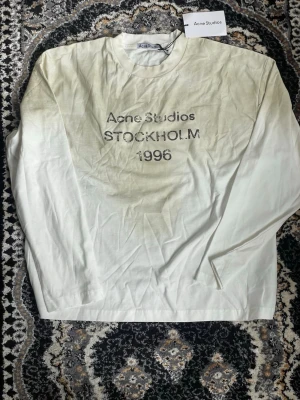 Vit långärmad topp Acne Studios - Vit långärmad topp från Acne Studios med tryck 'Acne Studios STOCKHOLM 1996' framtill. Klassisk rund halsringning och rak passform. Perfekt för dig som gillar minimalistisk och stilren design. Passar både till S/M