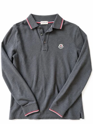 Mörkgrå Moncler långärmad piké - Snygg mörkgrå långärmad pikétröja från Moncler med klassisk krage och knäppning framtill. Tröjan har vita och röda detaljer på krage och ärmslut samt Moncler-logga på bröstet. Perfekt till jeans för en clean och stilren look. Jätte bra skick har endast ett litet litet hål längst ner på tröjan som man kan se på sista bilden 