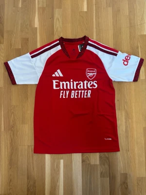 Arsenal matchtröja Saka 7 - Röd och vit Arsenal matchtröja med Saka 7 på ryggen. Tröjan har korta ärmar, rund hals och Adidas-logga på framsidan. Materialet är troligtvis polyester, perfekt för fotboll och träning. Emirates Fly Better tryck på bröstet och klubbmärke på vänster sida.