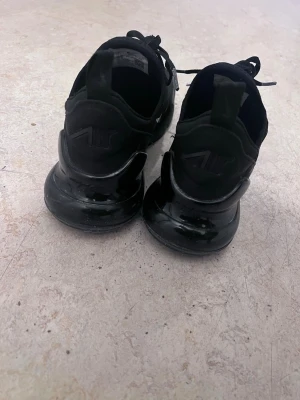 Nike Air Max 270 svarta sneakers - Säljer ett par helsvarta Nike Air Max 270 sneakers med tydlig Air-dämpning i sulan och klassisk Swoosh-logga på sidan. Skorna har mesh- och syntetmaterial för bra andningsförmåga och komfort. Snygg och sportig design med snörning och låg profil.