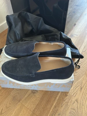 Mörkblå mocka loafers med vit sula - Snygga mörkblå loafers i mocka med stilren design och vit gummisula.  Köpta för 2000 kr knappt använda mer än 1 gång. Hälskyddet i skorna kan tas bort blev bara lagt till.  