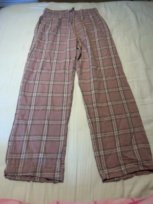 Rutiga rosa pyjamasbyxor - Mysiga pyjamasbyxor i rosa med rutigt mönster i vitt och grått. Byxorna har resår i midjan och dragsko för extra komfort. Perfekta för slappa dagar hemma eller en god natts sömn.