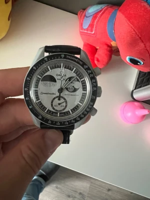 Speedmaster svart/silver klocka - Säljer en  Speedmaster klocka med svart läderarmband och silverfärgat boett. Urtavlan är vit med svarta detaljer och har flera subdials samt tachymeter på bezeln. Klassisk och sportig design som sticker ut på handleden. Ej äkta 