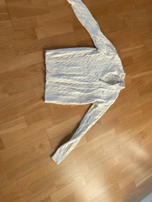Vit kabelstickad tröja - Säljer en vit kabelstickad tröja med v-ringning och långa ärmar från gant. Tröjan har ribbstickade muddar vid ärmslut och nederkant, vilket ger en snygg och klassisk look. Perfekt för dig som gillar stilrena och tidlösa plagg.