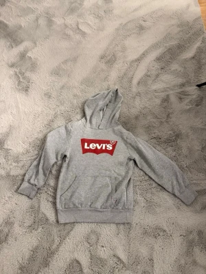 Grå Levi's hoodie med röd logga - Snygg grå hoodie från Levi's med klassisk röd logga på bröstet. Tröjan har huva, magficka och ribbade muddar vid ärmar och nederkant. Tillverkad i mjukt material som passar perfekt till en avslappnad stil.
