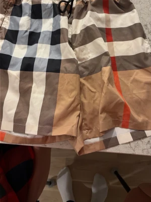 Burberry rutiga badbyxor - Säljer ett par klassiska badbyxor från Burberry med det ikoniska rutmönstret i beige, svart, vitt och rött. Badbyxorna har resår i midjan och innerbyxa i mesh för extra komfort. Perfekta för stranden eller poolen!