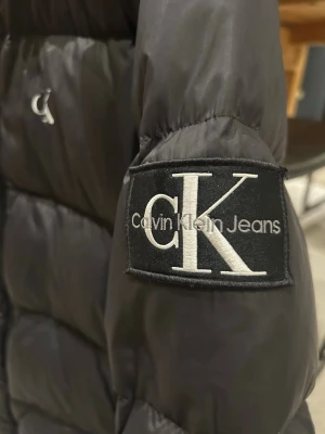 Svart pufferjacka från Calvin Klein - Säljer en svart pufferjacka från Calvin Klein Jeans med stor CK-logga på ärmen och mindre logga på bröstet. Jackan har dragkedja framtill, två sidofickor med dragkedja och hög krage. 