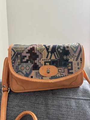 Axelväska med mönstrad textil och läder - Säljer en unik axelväska med beige läderdetaljer och lock i mönstrad textil i blå, grön och lila toner tillverkad på 90-talet.  Väskan har justerbar axelrem och ett litet metallmärke framtill. Perfekt för dig som gillar retro och boho-stil. Mått 28x18x10 cm 