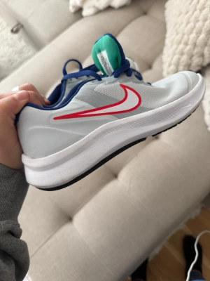 Nike vita sneakers med röd swoosh - Nike sneakers i vitt med röd swoosh på sidan, blå snören och detaljer samt grön plös. Ovandelen är i mesh för bra andningsförmåga och sulan är vit med svarta partier undertill. Perfekta för en sportig och avslappnad stil.