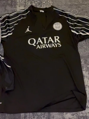 Svart PSG träningströja Jordan - Säljer en svart PSG träningströja från Jordan med vita detaljer och logga på bröstet. Tröjan har lång ärm, vita ränder på axlarna och Qatar Airways-tryck framtill. Perfekt för fotbollsträning eller chill.