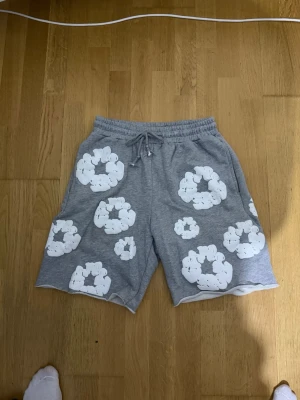 Grå shorts med vita blommor - Säljer ett par grå shorts med elastisk midja och snörning. Shortsen har ett unikt blommigt mönster i vitt över hela plagget och en bakficka. Perfekta för sommaren och riktigt sköna att bära.