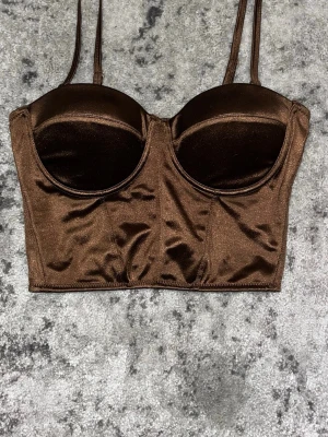 Brun korset topp  - Snygg brun bustier body med vadderade kupor och smala justerbara axelband. Tillverkad i glansigt material som ger en lyxig känsla. Perfekt att styla under en skjorta eller kavaj för en trendig look.