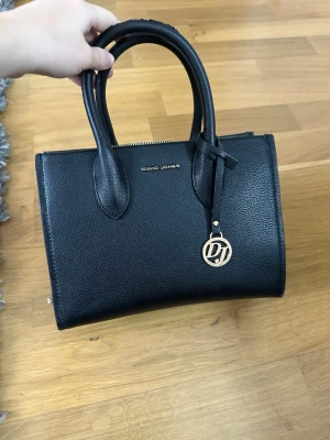 Svart handväska från David Jones - Stilren svart handväska från David Jones i konstläder med två rejäla handtag och guldfärgad logodetalj. Väskan har en klassisk, fyrkantig form och dragkedja upptill. Perfekt för dig som gillar enkel och tidlös design.