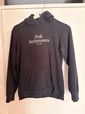 Svart hoodie Peak Performance - Säljer en svart hoodie från Peak Performance med vit logga och text 'Est. 1986' på bröstet. Hoodien har huva, känguruficka och ribbade muddar vid ärmslut och nederkant. Perfekt för en avslappnad och stilren look.