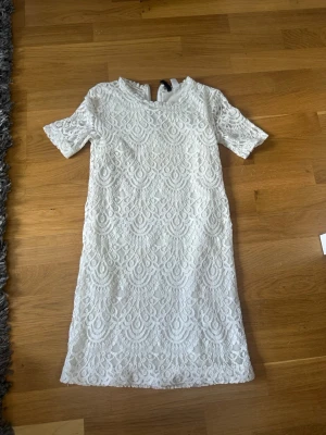 Vit spetsklänning med dragkedja - Säljer en elegant vit spetsklänning med korta ärmar och vackert broderat mönster. Klänningen har en smickrande rak passform och dragkedja i ryggen för enkel påklädning. Perfekt för sommarens alla tillfällen!