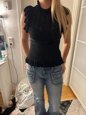 Svart blus med volangdetaljer - Säljer en svart blus med volangdetaljer vid ärmar och nederkant. Blusen har hög hals, är ärmlös med volang och ett diskret spetsmönster. Sitter tight i midjan och har en feminin siluett. Perfekt för dig som vill ha en chic och trendig topp.
