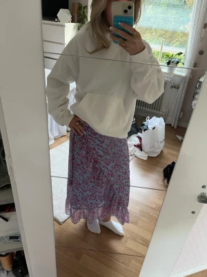 Blommig blå omlottkjol med volang - Säljer en ljusblå omlottkjol med rosa blommönster och volangkant. Kjolen är lång och har en luftig känsla, perfekt för vår och sommar. Materialet känns lätt och flowy, troligen polyester. Supersöt att styla med en enkel hoodie eller topp.