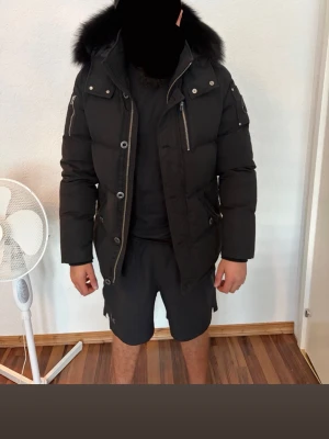 Svart moose knuckles  med 🦊 päls - Snygg svart dunjacka med stor pälskantad huva och flera dragkedjefickor. Jackan har en rak passform, tryckknappar och dragkedja framtill samt detaljer på ärmen. Perfekt för kalla vinterdagar och ger en stilren look. Kvitto finns