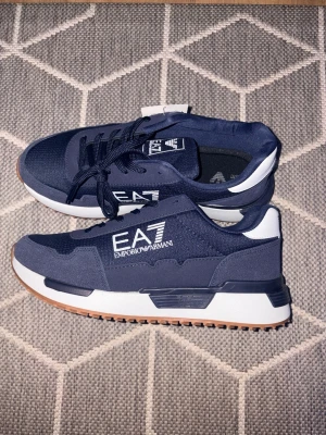 EA7 Emporio Armani mörkblå sneakers - Snygga mörkblå sneakers från EA7 Emporio Armani med vit logga på sidan, vita detaljer vid hälen och grov gummisula i brunt. Ovandelen är i textil och syntet, och skorna har klassisk snörning. Perfekta för dig som gillar stilrena och sportiga sneakers.