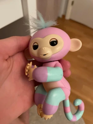 Fingerlings interaktiv apa rosa/turkos - Söt interaktiv Fingerlings apa i plast med rosa och turkos färg, beige ansikte och öron samt blått fluffigt hår på huvudet. Apan har böjbara armar och ben som kan greppa runt fingret och en randig svans. Den kan även prata och sättas på en penna!