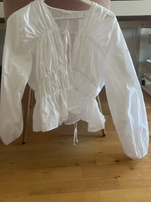 Vit blus med knyt och puffärm - Snygg vit blus med långa puffärmar och knytning fram. Blusen har en rynkad midja och v-ringad hals. Tillverkad i ett lätt och luftigt tyg som ger en avslappnad men trendig look. Perfekt att styla med jeans eller kjol för en chic outfit.