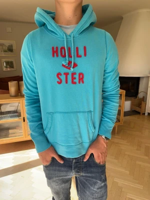 Hollister hoodie blå herr vintage - Snygg turkos Hollister hoodie med röd brodyr och klassisk logga framtill. Mjuk och bekväm modell med huva och magficka. Perfekt för en avslappnad preppy/surf-inspirerad stil.  #Hollister #hoodie #preppy #Y2K #surf #streetwear #college #vintage
