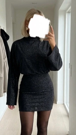 Svart glittrig kortklänning från Vila - Snygg svart kortklänning från Vila med glittrigt tyg och långärmad design. Klänningen har en hög hals och figurnära passform nedtill, perfekt för dig som vill sticka ut. Materialet känns mjukt och stretchigt, vilket gör den bekväm att bära.