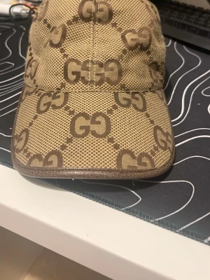 Beige keps med GG-mönster byter ellet säljer - Byter eller Säljer en beige keps från Gucci med det klassiska GG-monogrammet i brunt över hela kepsen. Kepsen har böjd skärm och justerbart spänne bak i brunt läder. Materialet är canvas med läderdetaljer för en lyxig känsla.
