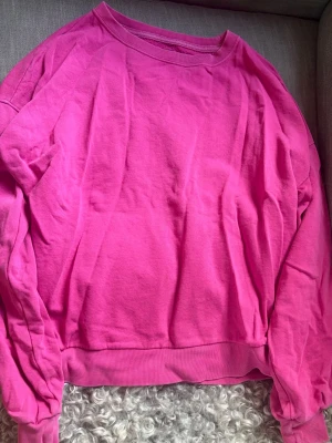 Klarrosa sweatshirt utan huva - En enkel och snygg klarrosa sweatshirt i mjukt bomullsmaterial. Tröjan har rund halsringning, långa ärmar och ribbade muddar vid ärmslut och nederkant. Perfekt för en avslappnad och färgstark look.