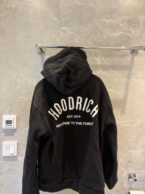 Svart hoodie från Hoodrich - Svart hoodie med dragkedja från Hoodrich. Stor logga och text 'WELCOME TO THE FAMILY' på ryggen samt mindre logga på bröstet. Utrustad med huva och snören, samt fickor framtill. Perfekt för en streetwear-look. Storlek M men S kan också ha den.